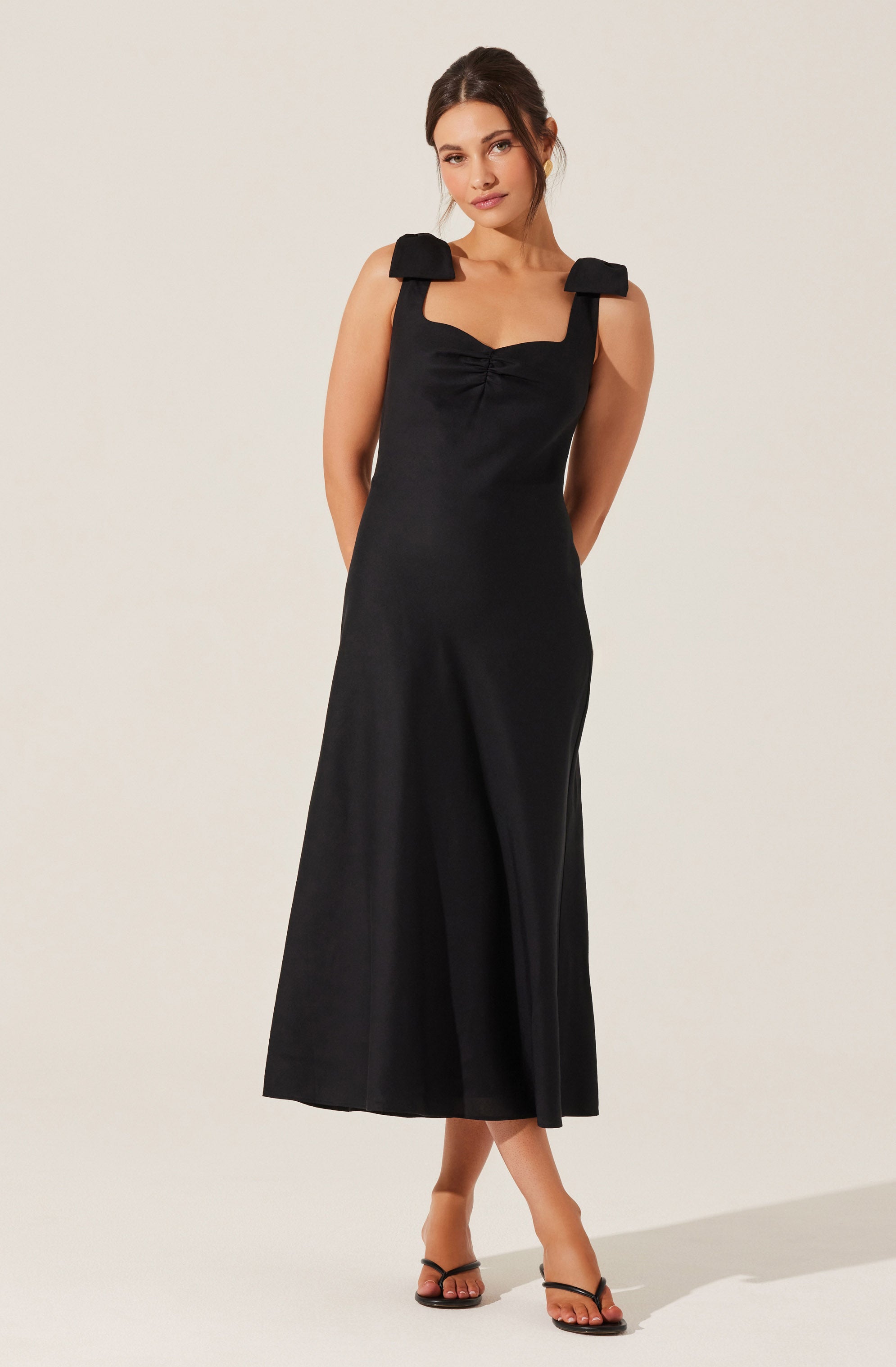 A woman is wearing a sleeveless black dress with wide shoulder straps that have large bow-like details on each shoulder. The dress has a fitted bodice with a gathered detail at the center of the neckline and a long, flowing skirt. She is also wearing black strappy high-heeled sandals.