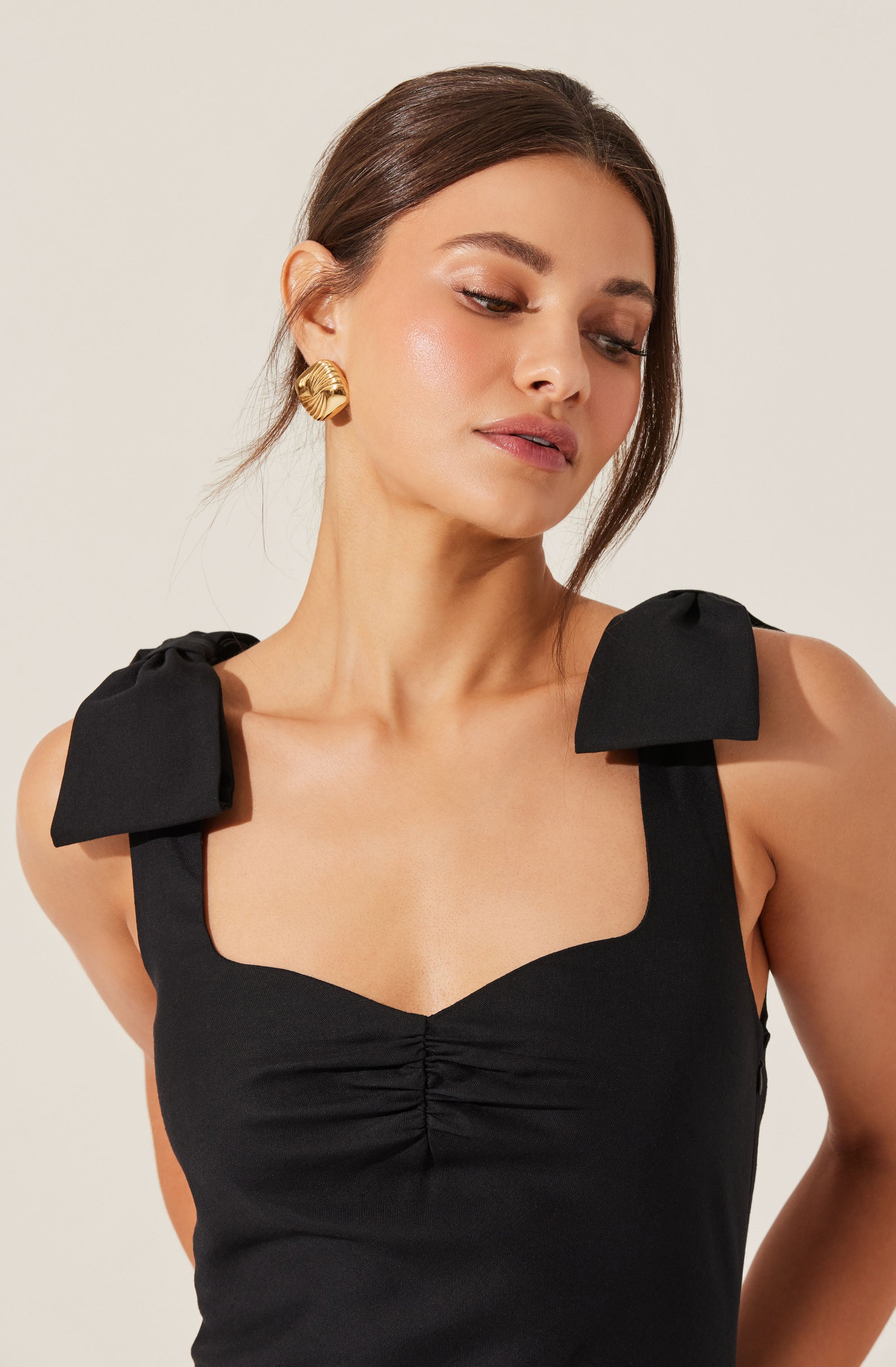 A woman is wearing a sleeveless black top with wide shoulder straps that have large bow details. The top has a fitted bodice with a gathered detail at the center of the bust. She is accessorized with large, gold, shell-shaped earrings.