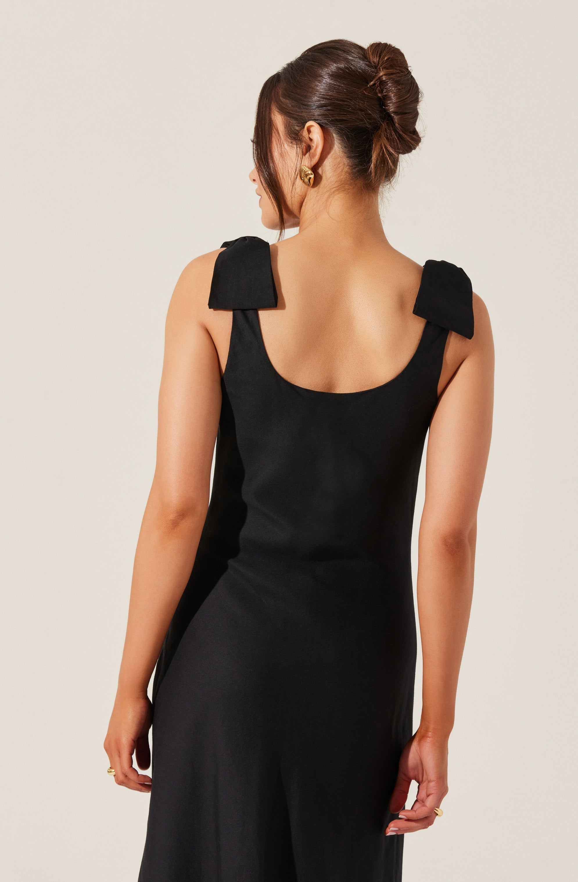 A woman is wearing a sleeveless black dress with wide shoulder straps that have a structured, bow-like design. The dress has a rounded neckline and a fitted silhouette. She is also wearing gold hoop earrings and a ring on her left hand. Her hair is styled in an elegant, low, voluminous bun.