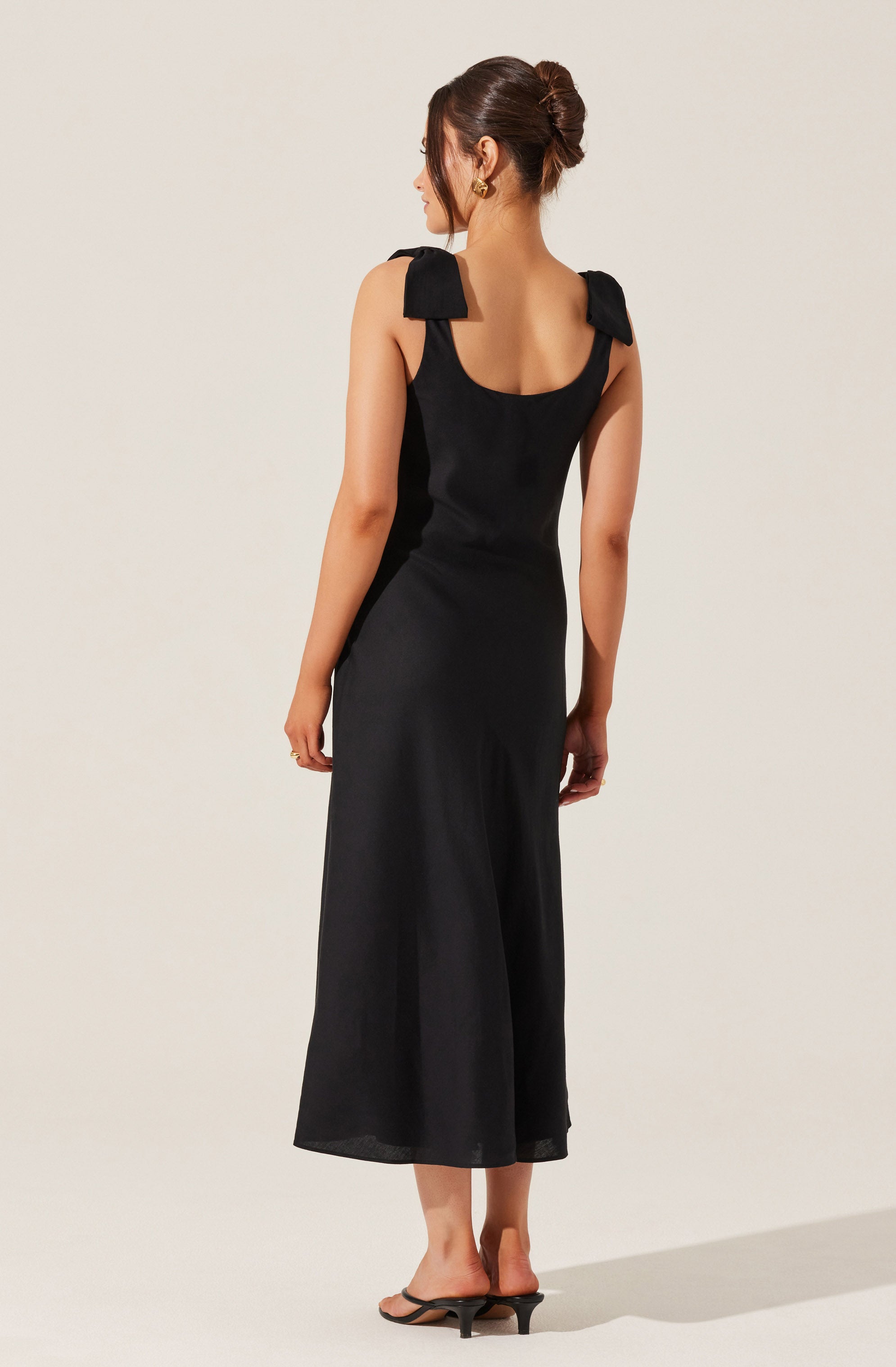 A woman is wearing a black sleeveless dress with shoulder ties that form bows. The dress has a fitted bodice and a flowing, wide-leg skirt that falls to mid-calf length. She is also wearing black open-toe sandals with a small heel.