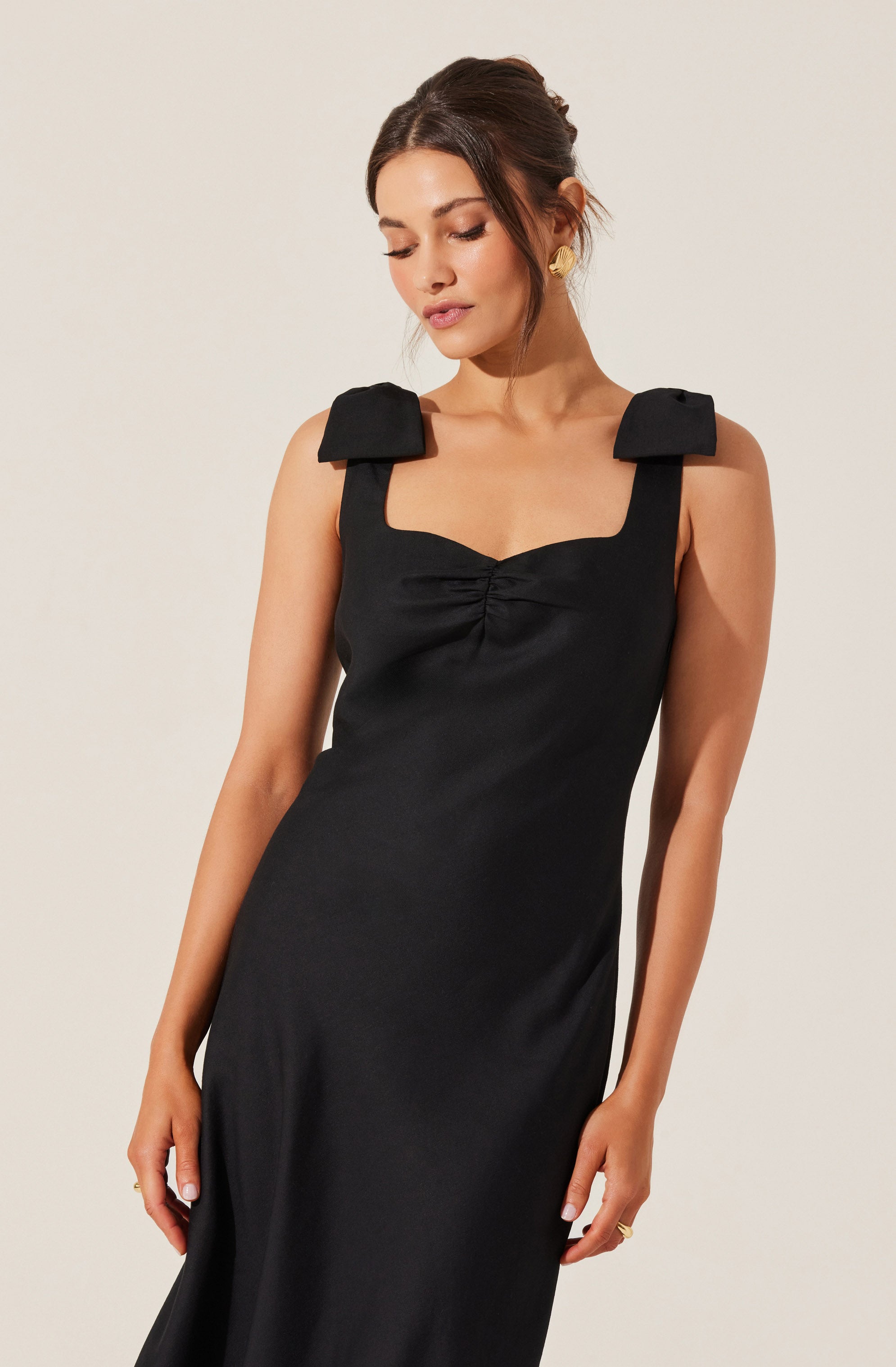 A woman is wearing a black sleeveless dress with wide shoulder straps that feature large bow details. The dress has a fitted bodice with a slight sweetheart neckline and a gathered detail at the center front. The fabric appears smooth and slightly shiny. She is accessorized with gold earrings and a ring, with her hair styled in an updo.