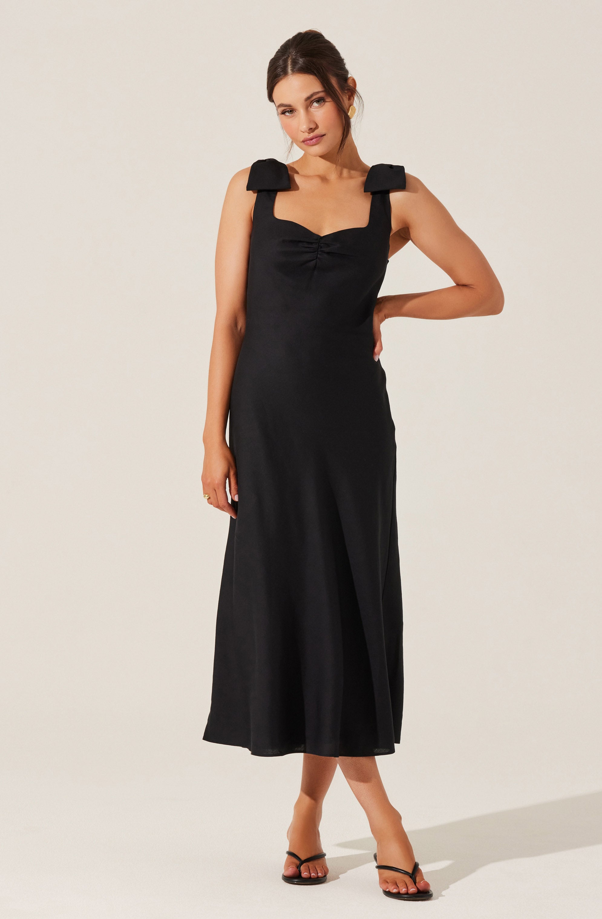 A woman is wearing a sleeveless black dress with wide shoulder straps. The dress features a sweetheart neckline with gathered fabric at the bust, and the length extends to her mid-calf. She has paired the dress with black open-toe heels.
