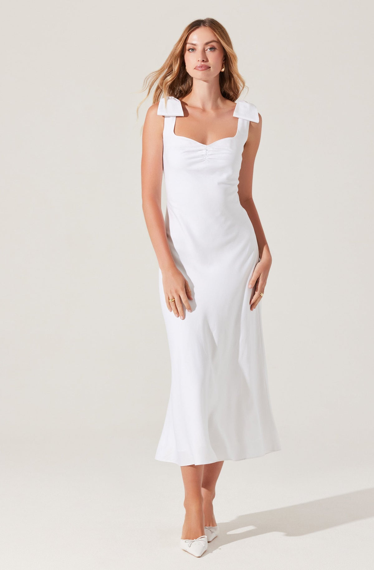 A woman is wearing a sleeveless white dress that has wide shoulder straps with prominent bow-like details on each shoulder, a fitted bodice with a sweetheart neckline, and a long, flowing skirt that reaches to her ankles. She is also wearing white pointed-toe shoes.