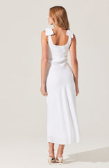A woman is wearing a long, elegant white dress with wide shoulder straps that have bow details on each shoulder. The dress has a fitted waist and flows down to the floor. She is also wearing white high-heeled shoes.