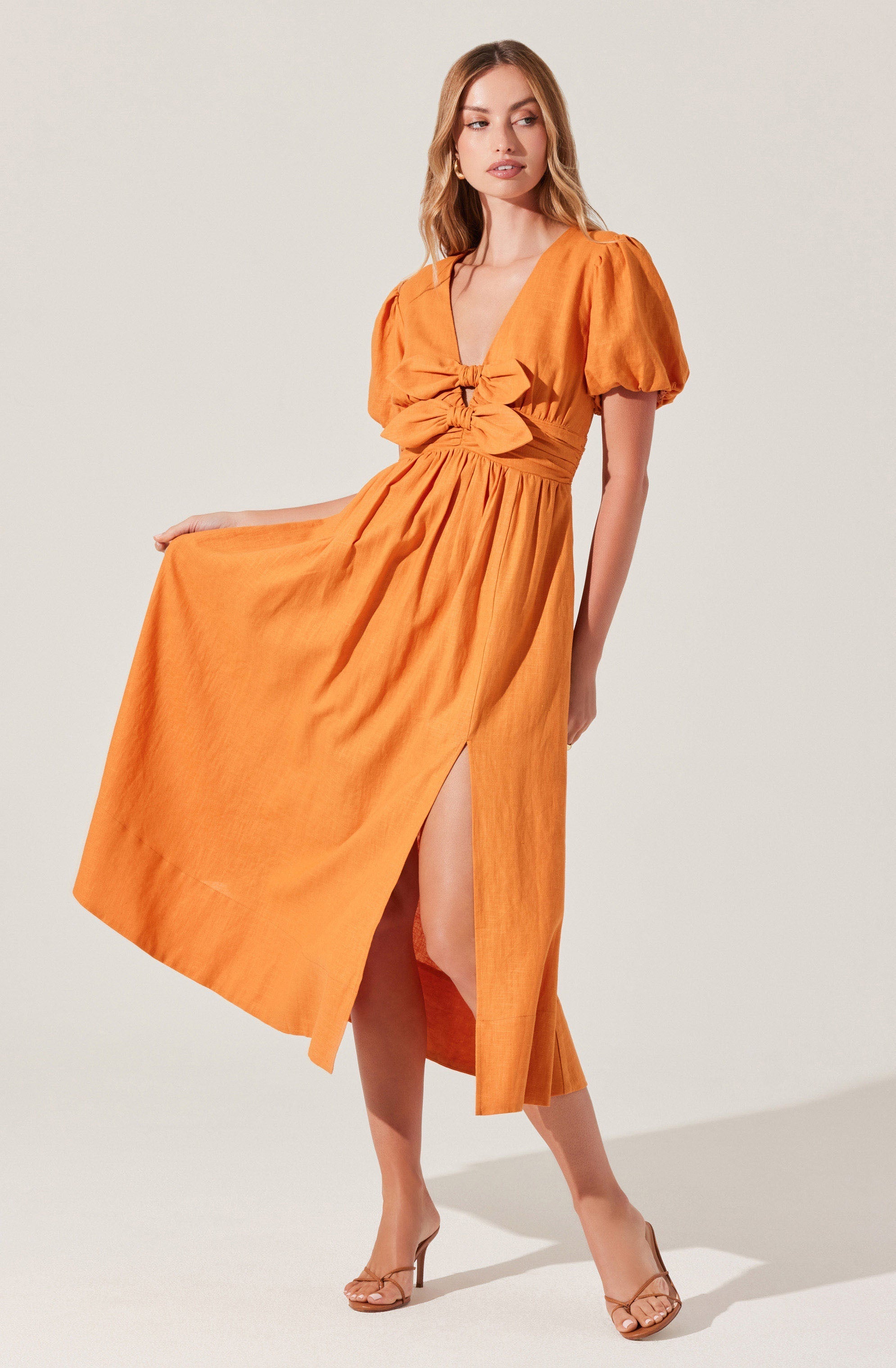 A woman is wearing a bright orange, midi-length dress with short, puffed sleeves and a deep V-neckline. The dress features a gathered detail at the bust with a large bow, and a slit on the side revealing her leg. She is also wearing nude high-heeled sandals.