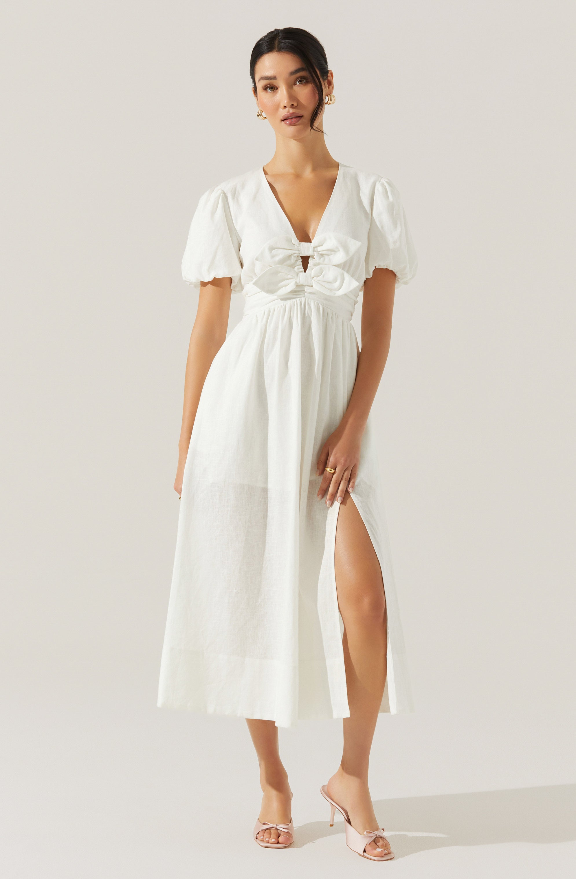 A woman is wearing a white maxi dress with short, puffed sleeves and a deep V neckline. The dress features a ruched bodice with a large bow at the front and a high slit on one side, revealing her leg. She is also wearing clear, open-toe high heels.