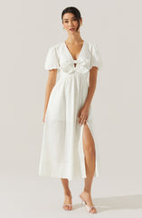 A woman is wearing a white maxi dress with short, puffed sleeves and a deep V neckline. The dress features a ruched bodice with a large bow at the front and a high slit on one side, revealing her leg. She is also wearing clear, open-toe high heels.