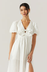 A woman is wearing a white dress with short, puffy sleeves and a deep V neckline. The bodice features a gathered design with a bow detail at the front, and the waistline is fitted. The skirt is long and flowing with a side slit.