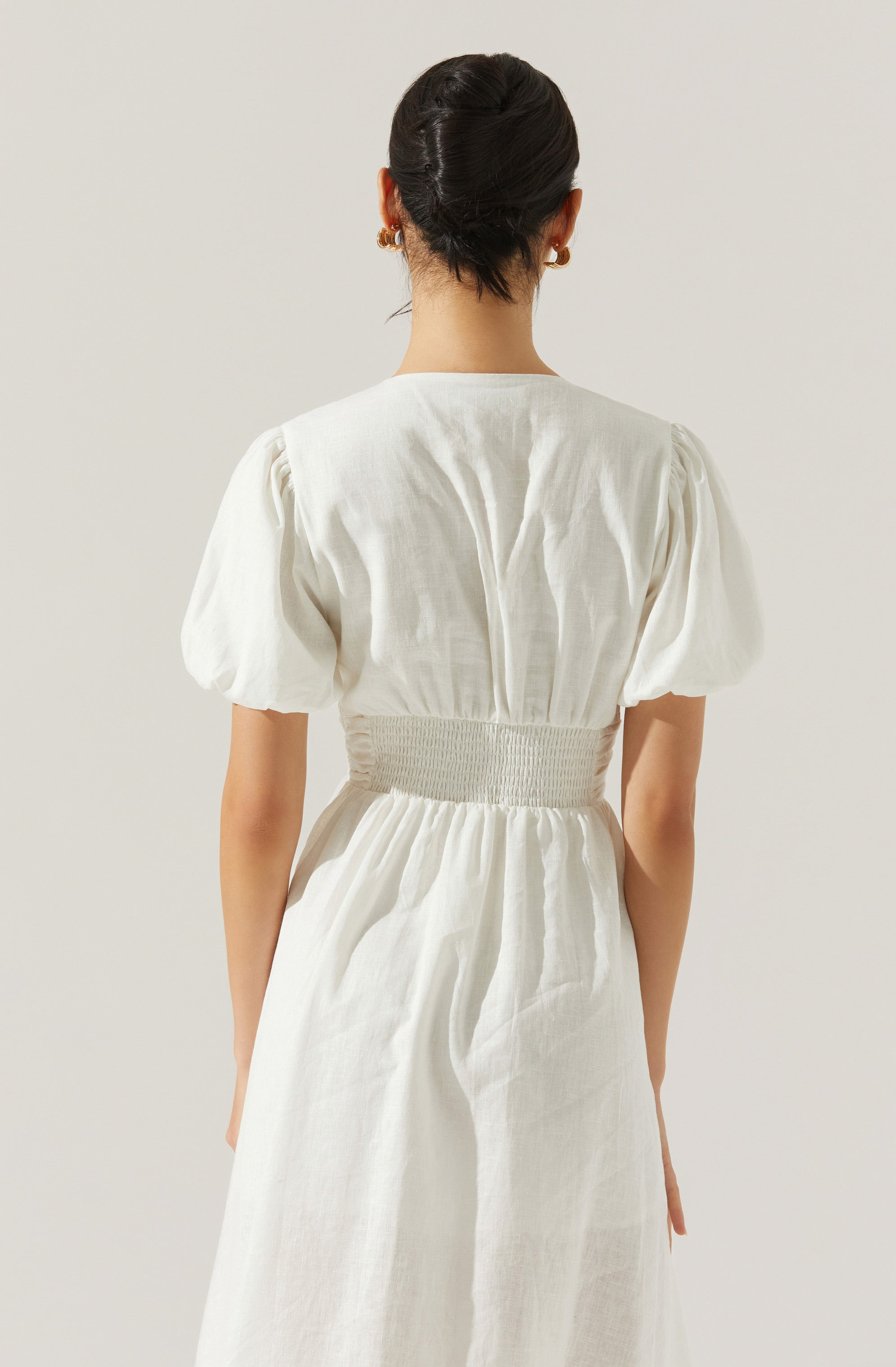 A woman is wearing a white dress with puffed short sleeves, a fitted waistband, and a gathered skirt. The dress appears to be made of a light, airy fabric, and the back features a central seam running vertically. The waistband is wide and elastic, creating a cinched waist effect. The woman has dark hair styled in an elegant updo and is accessorized with earrings.