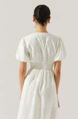 A woman is wearing a white dress with puffed short sleeves, a fitted waistband, and a gathered skirt. The dress appears to be made of a light, airy fabric, and the back features a central seam running vertically. The waistband is wide and elastic, creating a cinched waist effect. The woman has dark hair styled in an elegant updo and is accessorized with earrings.
