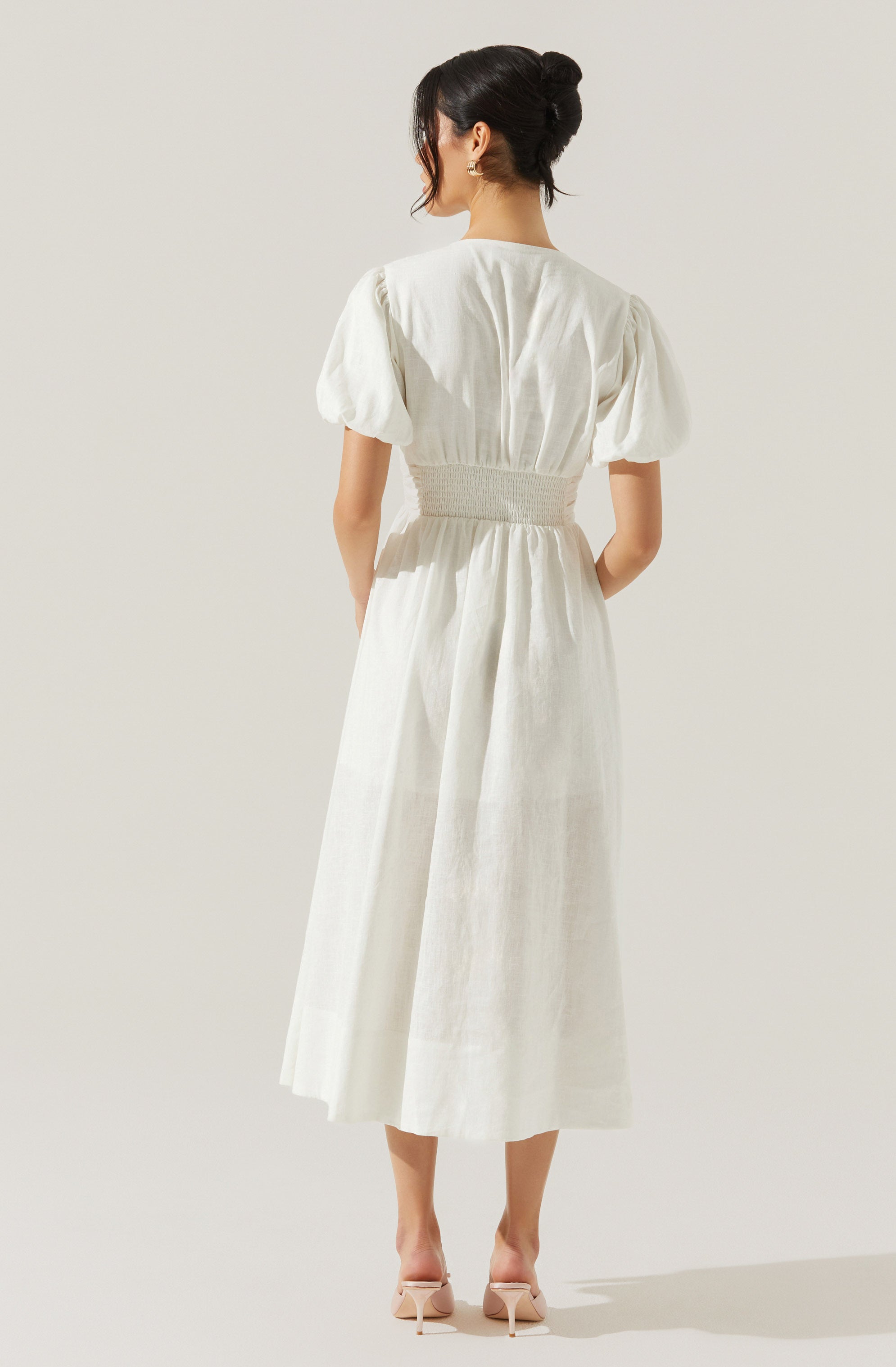 A woman is wearing a long, white dress with puffed short sleeves, a fitted waist with gathered detailing, and a full-length skirt that reaches mid-calf. She is standing with her back to the camera, and her hair is styled in an elegant updo. She is also wearing light-colored, open-toe high heels.