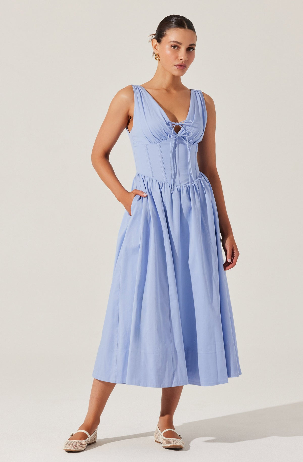 A woman is wearing a light blue sleeveless dress with a fitted bodice featuring a lace-up detail at the front. The dress has a full, gathered skirt that reaches mid-calf length. She completes the look with white, closed-toe shoes that have a small heel.