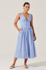 A woman is wearing a light blue sleeveless dress with a fitted bodice featuring a lace-up detail at the front. The dress has a full, gathered skirt that reaches mid-calf length. She completes the look with white, closed-toe shoes that have a small heel.