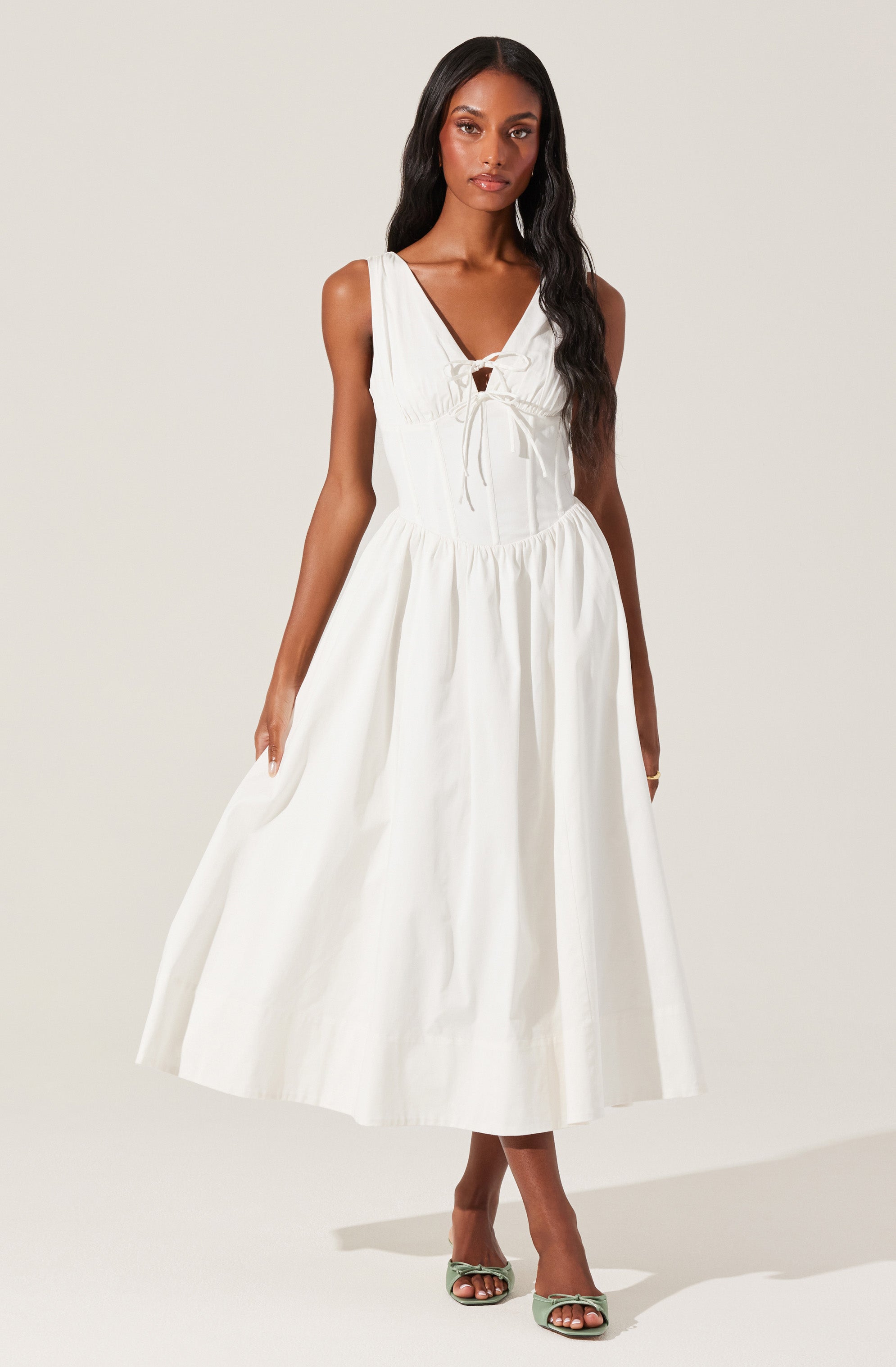A woman is wearing a sleeveless, white, midi-length dress with a gathered waist. The dress features a V-neckline with a small tie detail at the front. She is also wearing light green open-toe heels.