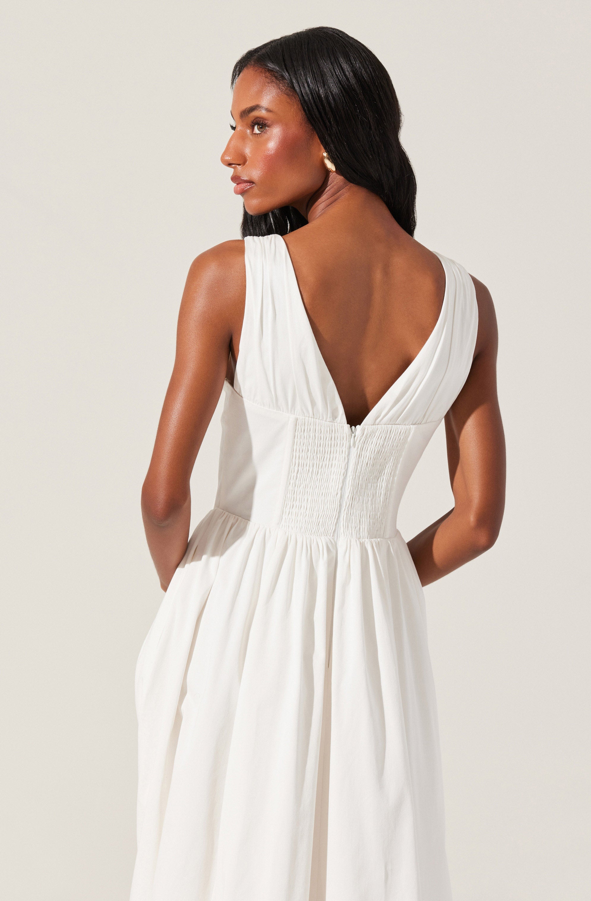 A woman is wearing a sleeveless white dress with a deep V back. The top of the dress has gathered fabric at the shoulders, and the waist features smocking detail. The skirt of the dress is loose and flowing, with pleats creating a full silhouette. The fabric appears lightweight and airy.