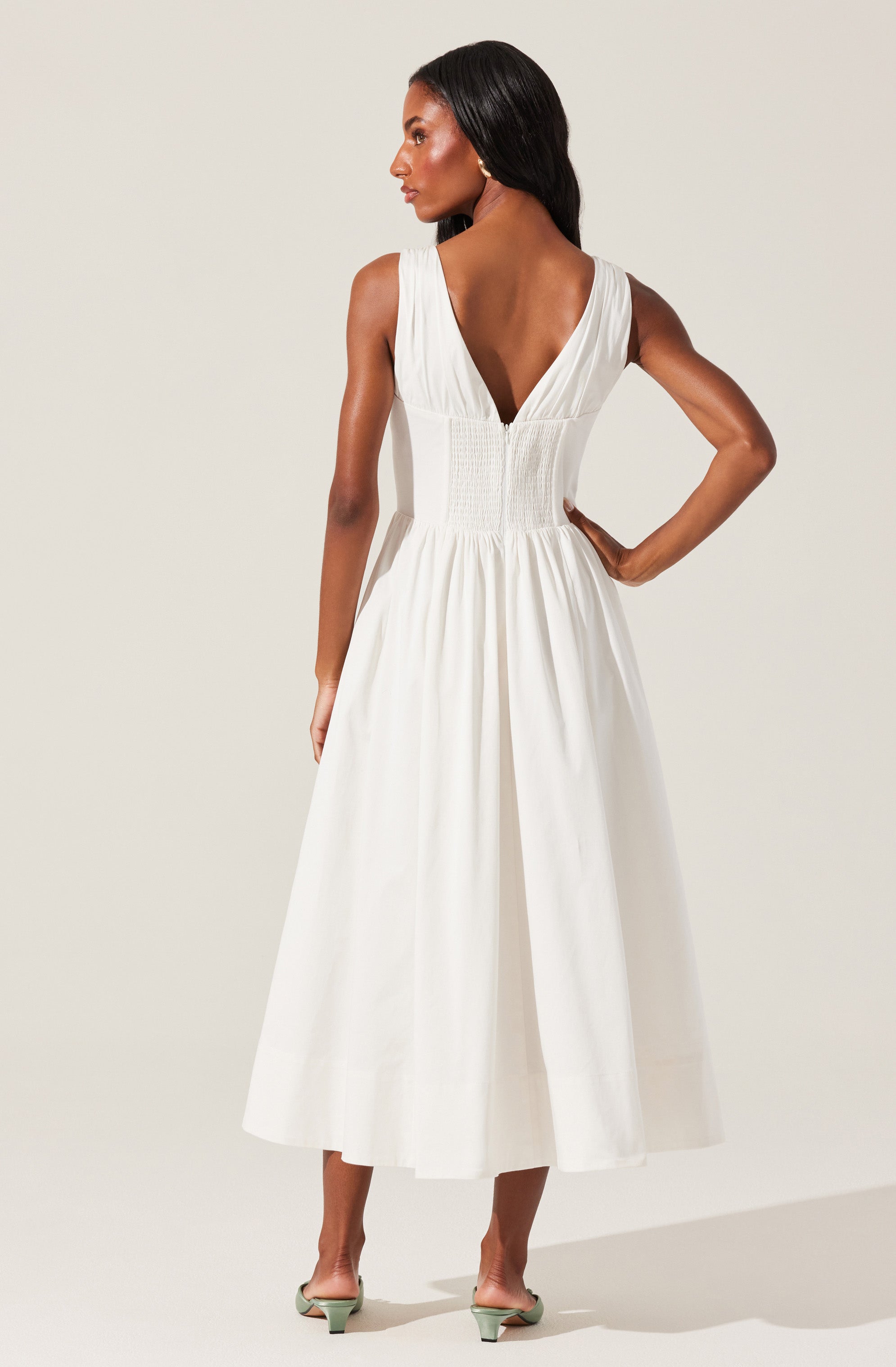 A woman is wearing a sleeveless, ankle-length white dress with a deep V-back. The dress features a fitted, smocked bodice and a gathered, flowing skirt. She is styled with open-toed heels and has long, straight black hair.