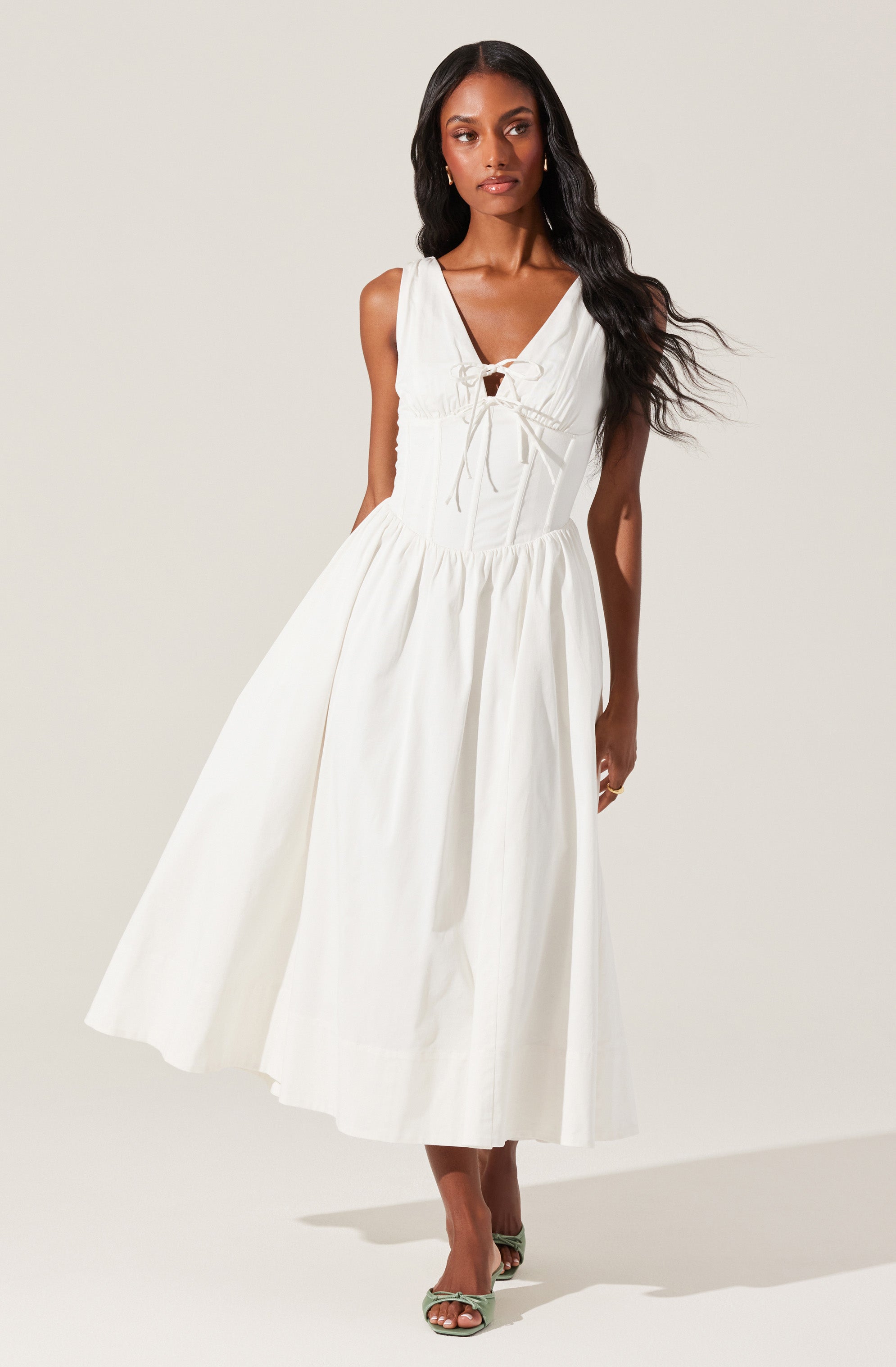 A woman is wearing a sleeveless, white, ankle-length dress with a fitted bodice featuring a lace-up detail at the front. The dress has a full, flowing skirt and a V-shaped neckline. She is also wearing open-toe high-heeled shoes in a light color.