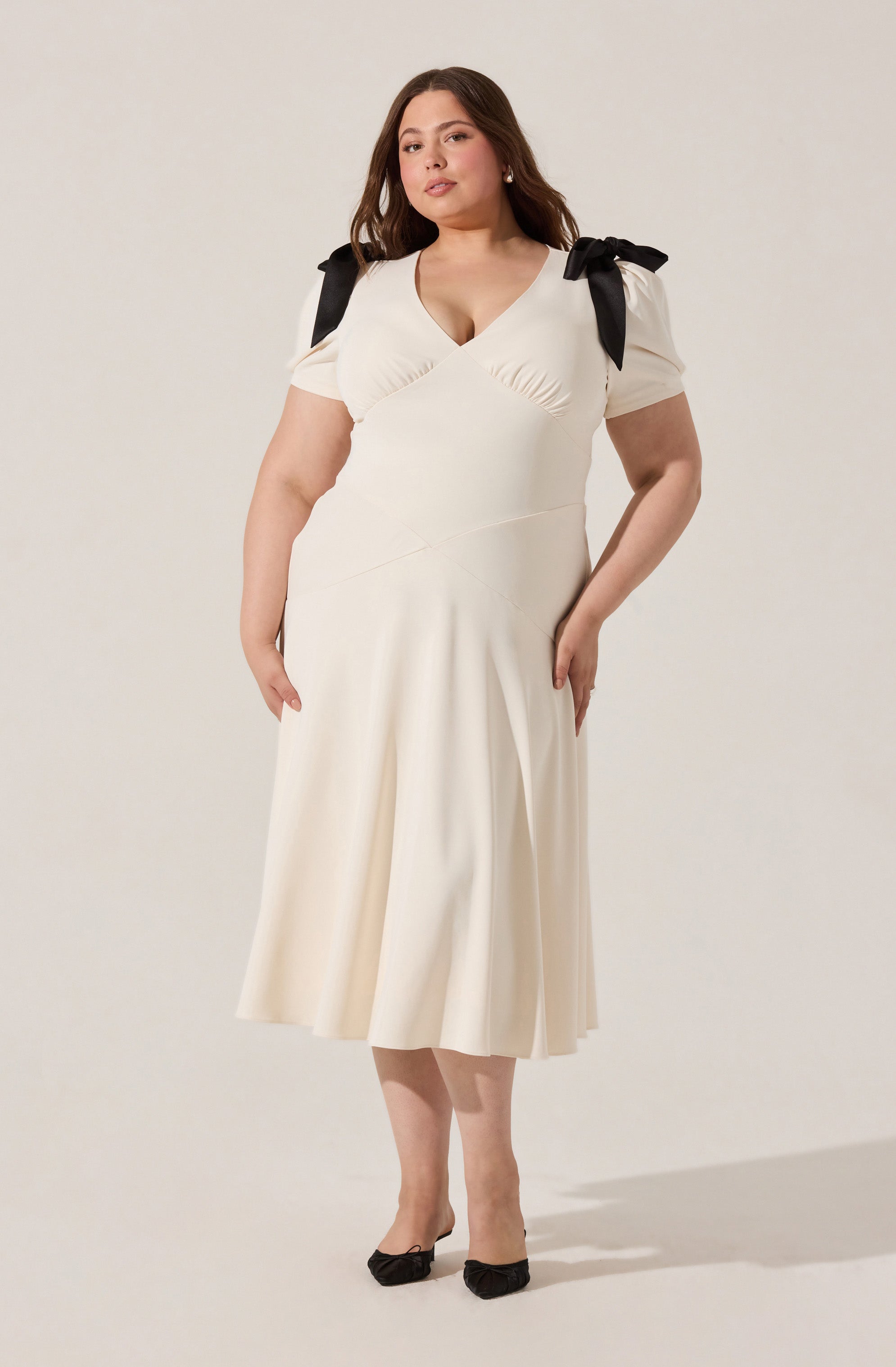 A woman is wearing a cream-colored, midi-length dress with a fitted bodice and a flared skirt. The dress features short puff sleeves with black ribbon bows on each shoulder. The neckline is a V-shape with subtle ruching details.