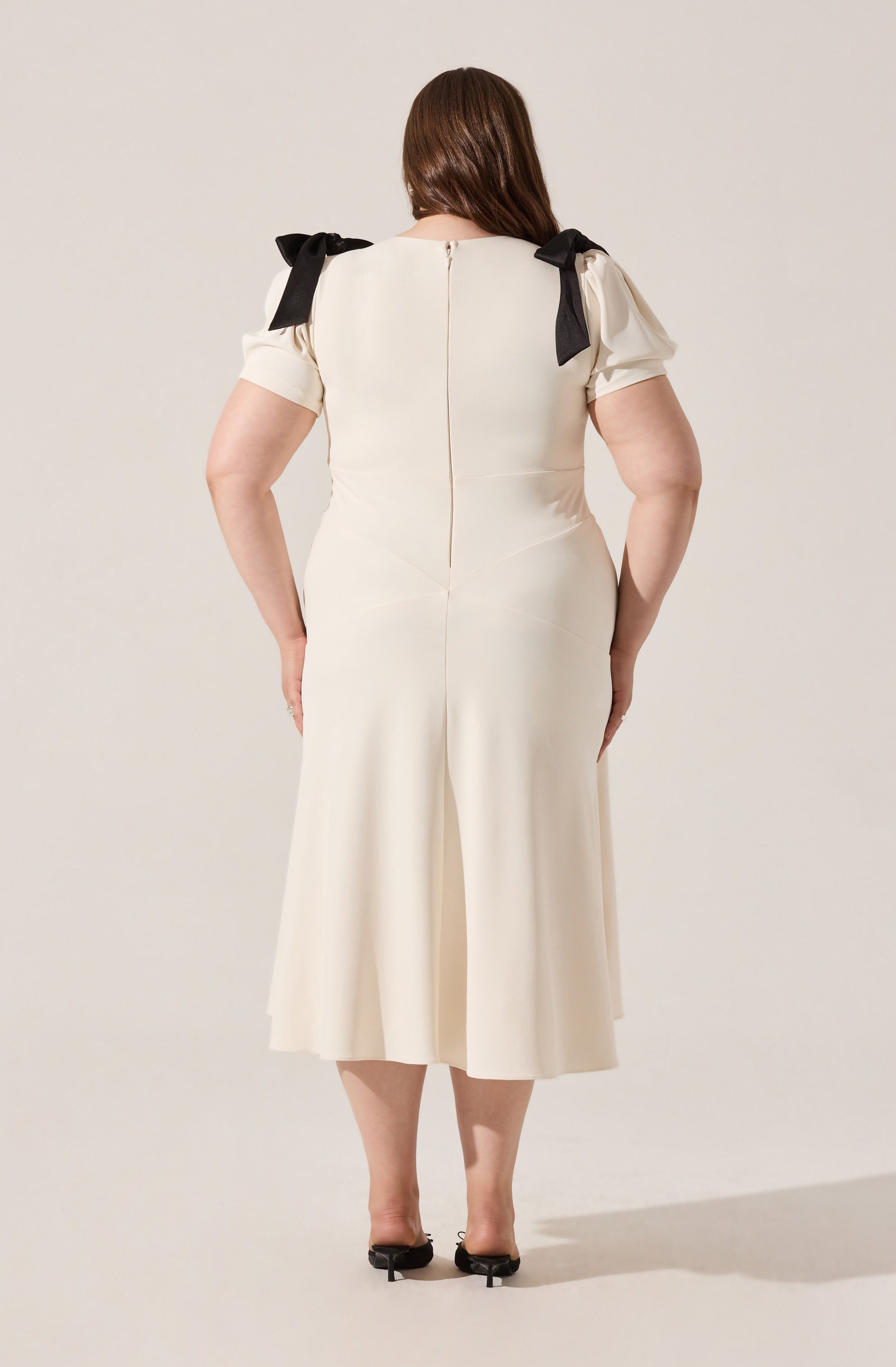 A woman is wearing a cream-colored dress with short puffed sleeves that have black bows on the shoulders. The dress has a fitted waist and a back zipper, with pleats in the skirt that fall to mid-calf length. She is wearing black open-toe heels.