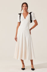 The dress is a white, midi-length gown with a deep V neckline. It features puffed short sleeves with black bows on each shoulder. The fabric appears smooth and flowing, and the skirt has pleats that create a full, elegant silhouette. The outfit is paired with black pointed-toe shoes.