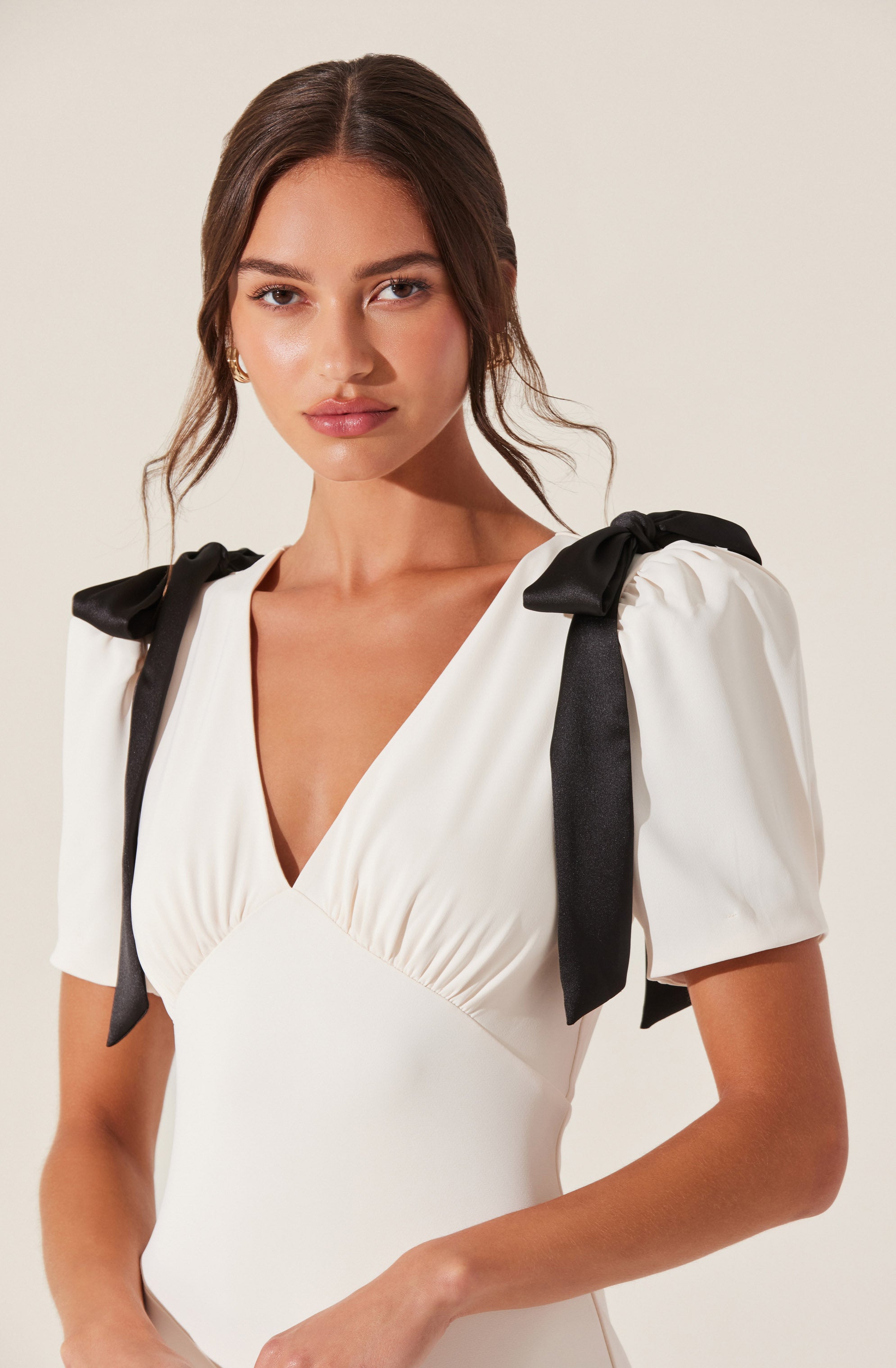 The woman is wearing a white dress with a deep V neckline and short puffed sleeves. The dress features black ribbon shoulder ties that are tied into bows, adding a decorative element to the shoulders.