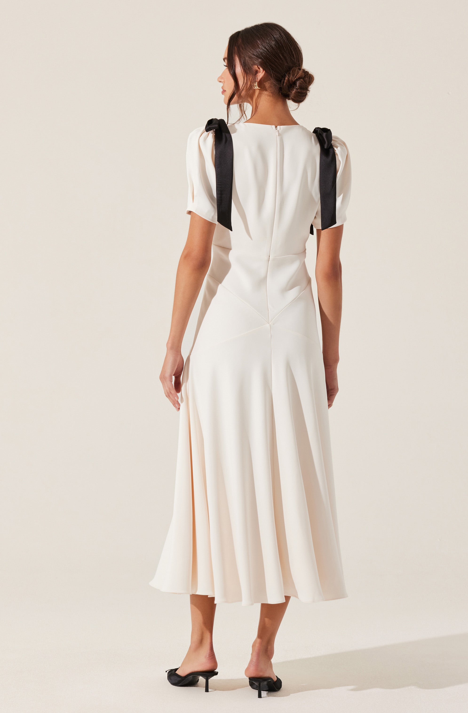 A woman is wearing an elegant, white dress that features a fitted bodice and a flowing, pleated skirt reaching mid-calf. The dress has short, puffed sleeves and black ribbon shoulder details tied into bows. She is also wearing black, open-toe, low-heeled shoes.