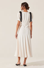 A woman is wearing an elegant, white dress that features a fitted bodice and a flowing, pleated skirt reaching mid-calf. The dress has short, puffed sleeves and black ribbon shoulder details tied into bows. She is also wearing black, open-toe, low-heeled shoes.
