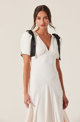 A woman is wearing a white dress with a deep V neckline and short puffed sleeves. The dress features black ribbon shoulder ties that create a bow on each shoulder. The fabric appears smooth and elegant, with subtle stitching details on the skirt.