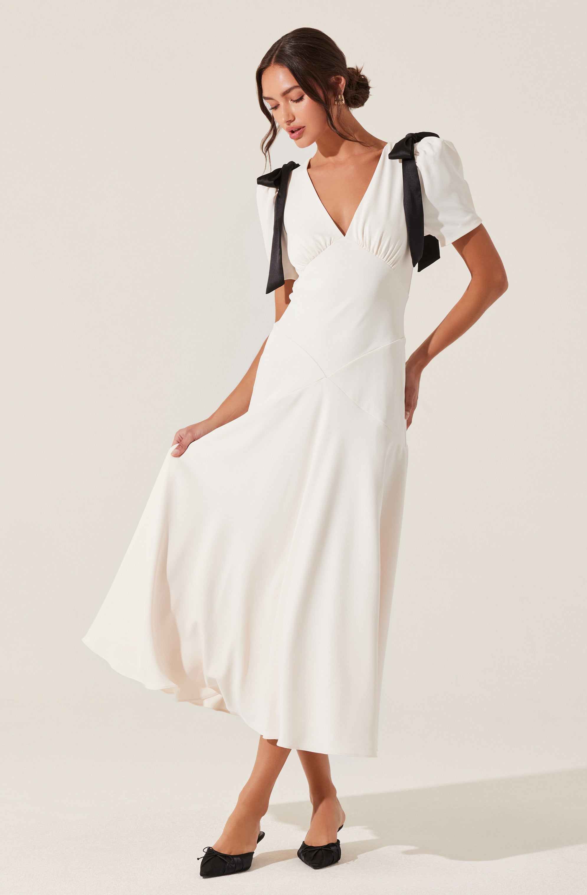 A woman is wearing a white, ankle-length dress with a V-neckline. The dress has puffed short sleeves with black bows on the shoulders. She is also wearing black pointed-toe shoes with small bows on the front.