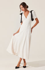 A woman is wearing a white, ankle-length dress with a V-neckline. The dress has puffed short sleeves with black bows on the shoulders. She is also wearing black pointed-toe shoes with small bows on the front.