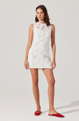 A young woman is wearing a sleeveless, white, knee-length dress with a crochet or lace pattern featuring geometric shapes and openwork details. She has on red flat shoes with a small bow on each.