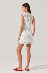 A woman is wearing a sleeveless, white lace dress with intricate geometric and floral patterns. The dress is knee-length with a scalloped hem and appears lightweight, suitable for warm weather. She is also wearing red slip-on flats and gold hoop earrings.