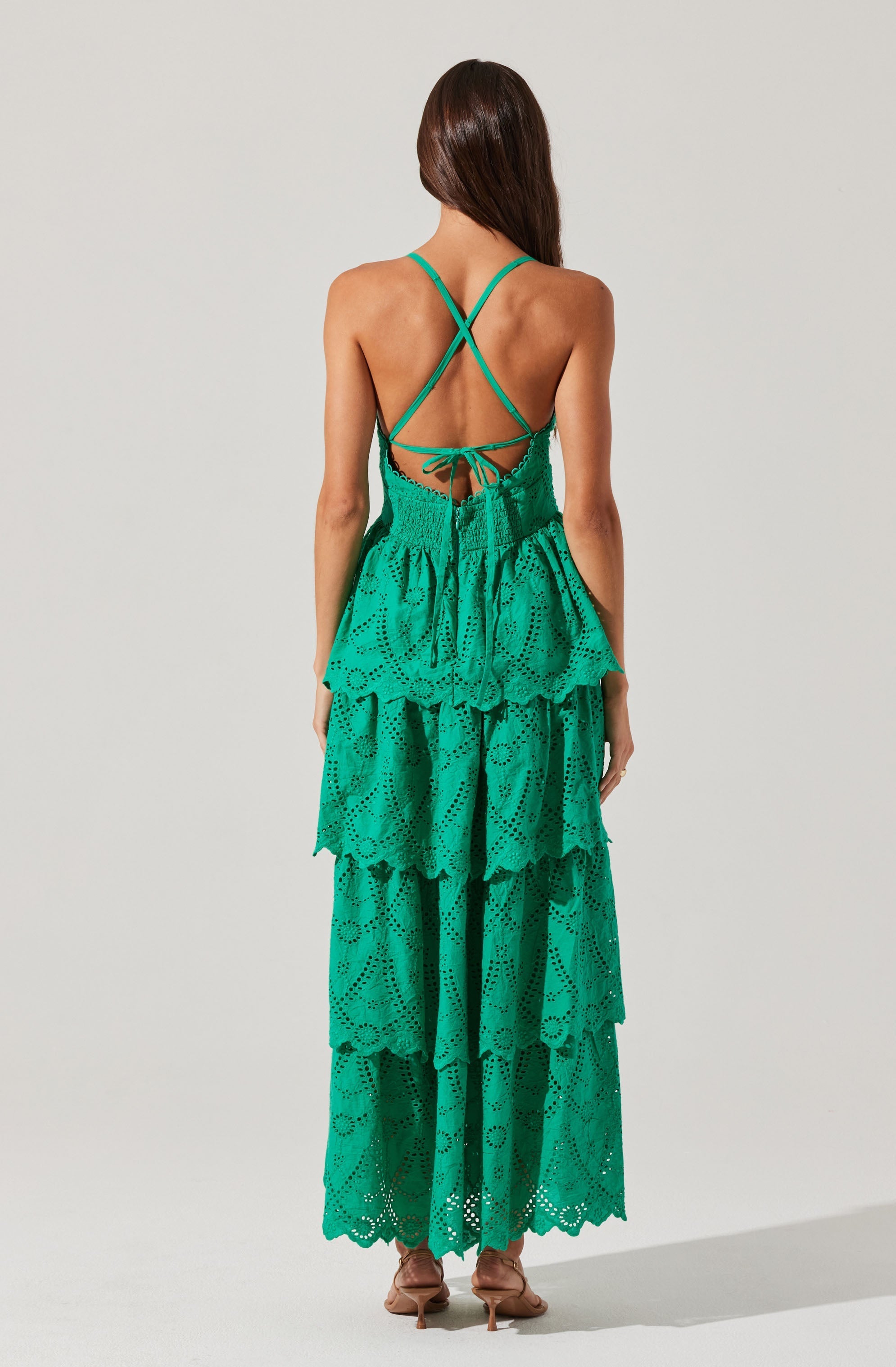 A woman is wearing a long, flowing, green dress with multiple tiers. The dress features eyelet embroidery and ruffled layers throughout the length. The back of the dress has thin, crisscrossing spaghetti straps that tie at the waist, revealing her back.