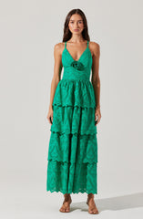 A woman is wearing a long, bright turquoise dress made of eyelet fabric with intricate cut-out patterns. The dress features a fitted bodice with adjustable spaghetti straps, a decorative rosette detail at the center of the bust, and a tiered, flowing skirt with multiple ruffled layers that extend to the floor.