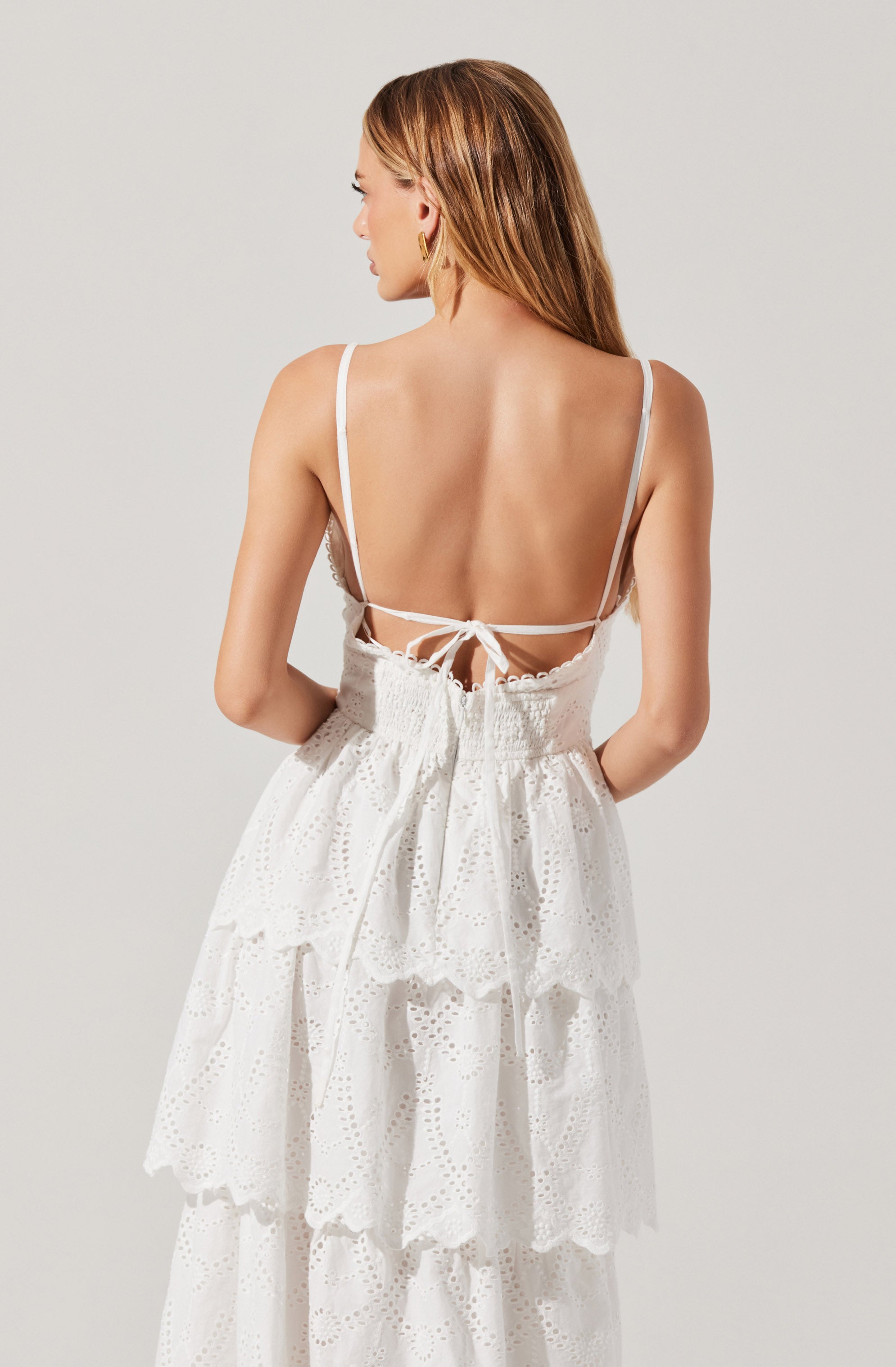 A woman is wearing a white, backless dress with thin spaghetti straps. The dress features a tiered design with eyelet embroidery on the fabric, creating a ruffled and textured appearance. The back of the dress has a tie closure at the waist.