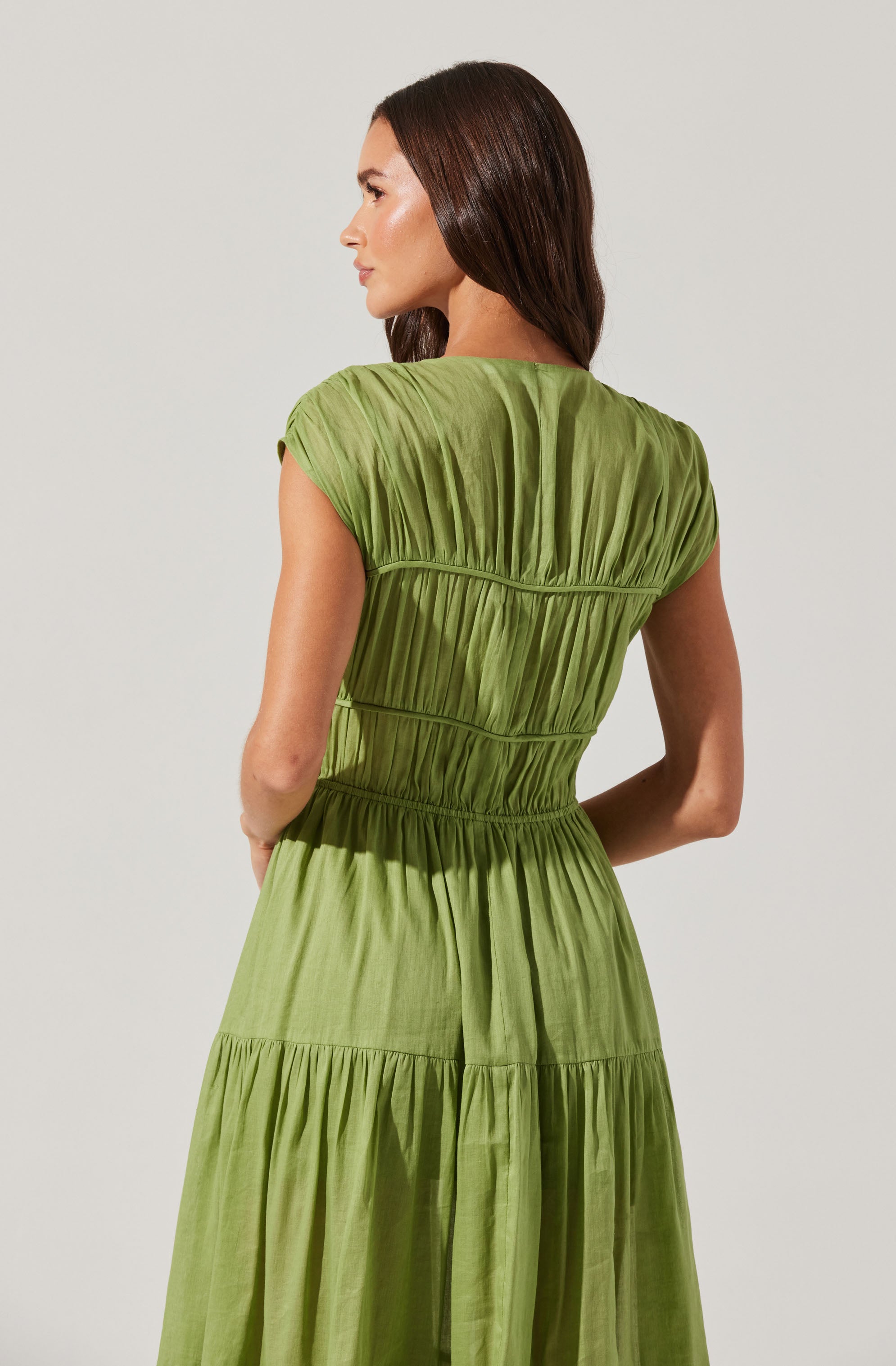 A woman is wearing a light green, tiered dress made of a soft, flowing fabric. The dress features short, gathered sleeves and multiple horizontal seams that create a gathered, textured effect throughout the garment. The waistline is slightly cinched, and the skirt section is gathered with a tiered design.