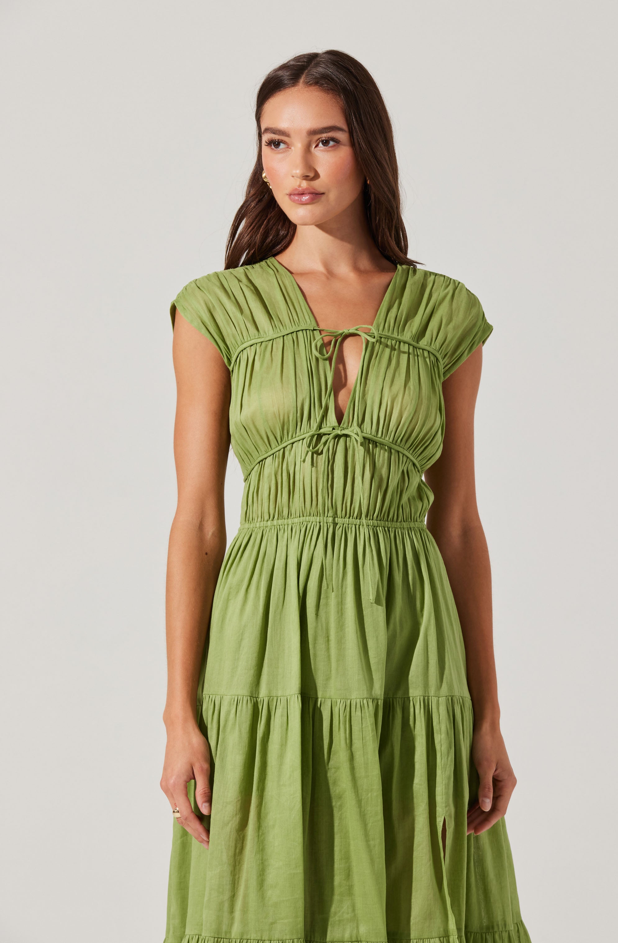 A woman is wearing a sleeveless, light green dress made of a lightweight, slightly shiny fabric. The dress features gathered details at the shoulders, a deep V-neckline with two ties at the front, a fitted waist with ruched elastic, and a tiered skirt with gathered sections creating a flowing silhouette.