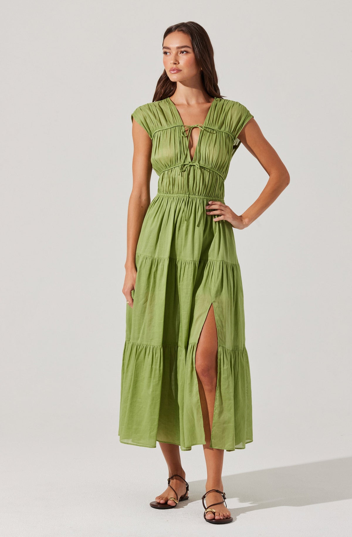 A woman is wearing a long, lime green dress with gathered details and a tiered skirt. The dress features a deep V neckline with a small keyhole and ties at the bust. It has short, cap sleeves and a high waistline. The skirt has a slit up to the knee, revealing her leg. She is also wearing strappy, flat sandals.
