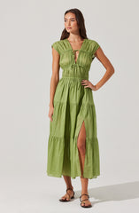 A woman is wearing a long, lime green dress with gathered details and a tiered skirt. The dress features a deep V neckline with a small keyhole and ties at the bust. It has short, cap sleeves and a high waistline. The skirt has a slit up to the knee, revealing her leg. She is also wearing strappy, flat sandals.