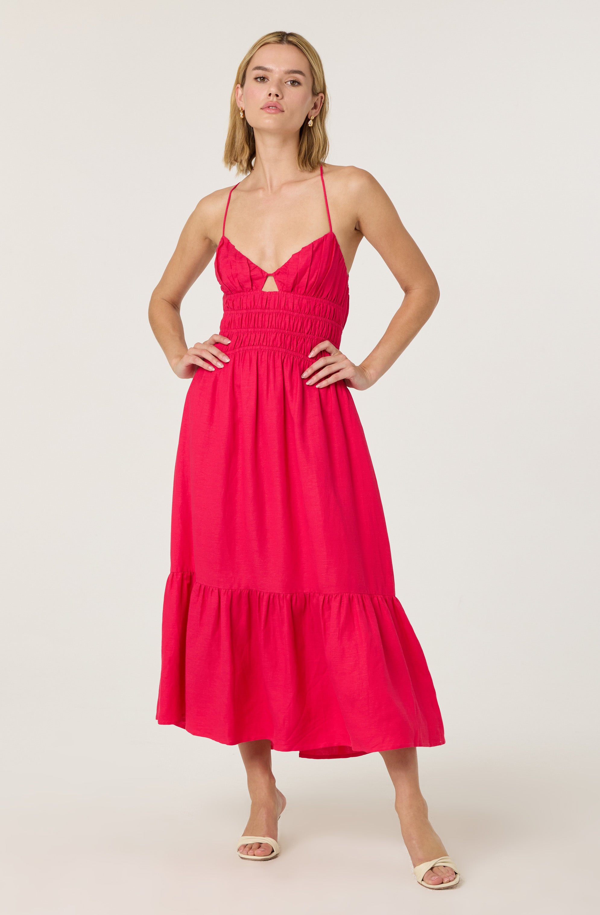 A woman is wearing a bright pink maxi dress with thin spaghetti straps. The dress has a fitted, ruched bodice with a small cutout detail at the center and a loose, gathered skirt that reaches the ankles. She is standing with her hands on her hips.