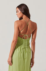 A woman is wearing a sleeveless dress with thin, crisscrossing spaghetti straps in a light green color. The top portion of the dress is ruched with elastic, and it has a drawstring at the waist with ties at the back. The skirt portion is flowy and gathered at the waist.