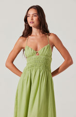 A woman is wearing a light green sundress with thin spaghetti straps. The top of the dress features gathered fabric and a small keyhole cutout below the bust. The waistline appears to be elasticized with horizontal ruched detailing, and the skirt is long and flowing with a gathered, full silhouette.