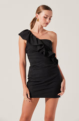 A woman is wearing a black, one-shoulder mini dress with ruffle details along the strap and down the front. The dress is fitted and has a slightly gathered design around the waist and hips. She accessorizes with gold earrings and rings.