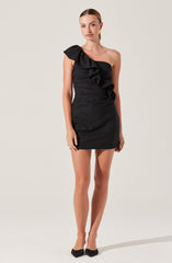 A woman is wearing a black, one-shoulder dress featuring a large ruffle detail that runs from the shoulder down the front. The dress is fitted and short in length. She has paired the dress with black shoes with a bow on top.