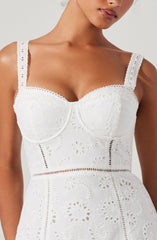 A woman is wearing a white sleeveless dress with embroidered eyelet patterns. The dress features wide shoulder straps, a fitted bodice with visible boning, and intricate cut-out embroidery forming floral and geometric designs throughout the fabric.