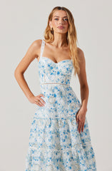 A woman is wearing a sleeveless, white dress with intricate blue floral and lace embroidery. The dress features a fitted bodice with bustier-style cups and wide shoulder straps, and a tiered, flowing skirt.