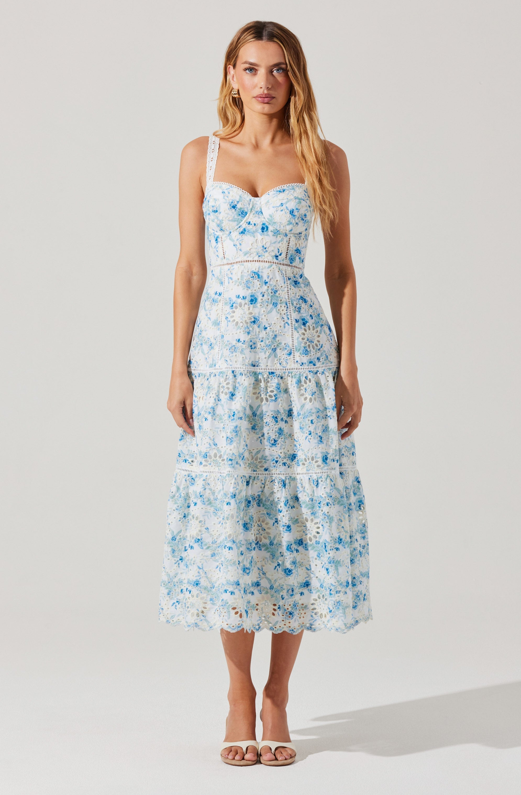 A woman is wearing a sleeveless, ankle-length dress with a fitted bodice and a full skirt. The dress features a white base with a blue floral pattern throughout, including on the wide shoulder straps, bodice, and skirt. The fabric appears lightweight and textured, with a tiered design on the skirt and delicate lace or embroidery details. She is also wearing nude-colored open-toe sandals.