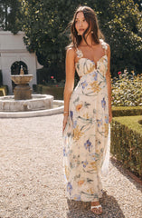 A woman is wearing a long, sleeveless dress with a floral pattern. The dress features a sweetheart neckline with decorative flowers on the straps and a flowy, sheer fabric that reaches the ground. The floral pattern includes purple, yellow, and orange flowers with green leaves on a light, possibly cream or white background. She is also wearing large, round earrings, a bracelet, and white open-toe sandals.