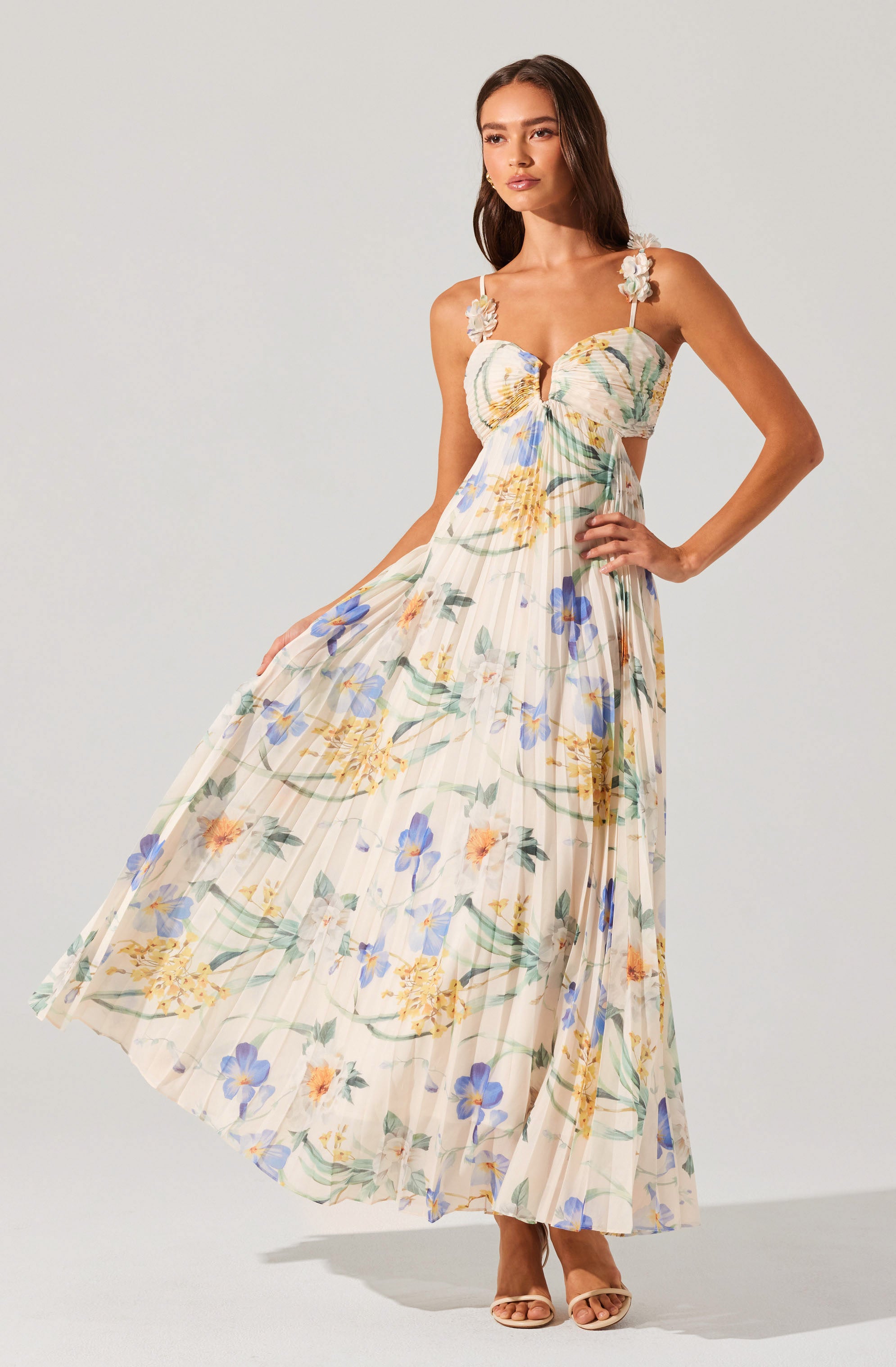 A woman is wearing a sleeveless, ankle-length dress with a floral print in shades of yellow, blue, orange, and green on a light, cream-colored background. The dress features ruffled, adjustable shoulder straps with floral accents, a fitted bodice with a sweetheart neckline, and a flowing pleated skirt. She is also wearing open-toed, nude-colored high heels.