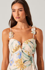 A woman is wearing a white dress with a floral pattern featuring blue, yellow, and green colors. The dress has ruched detailing at the bust and thin shoulder straps decorated with white fabric flowers.
